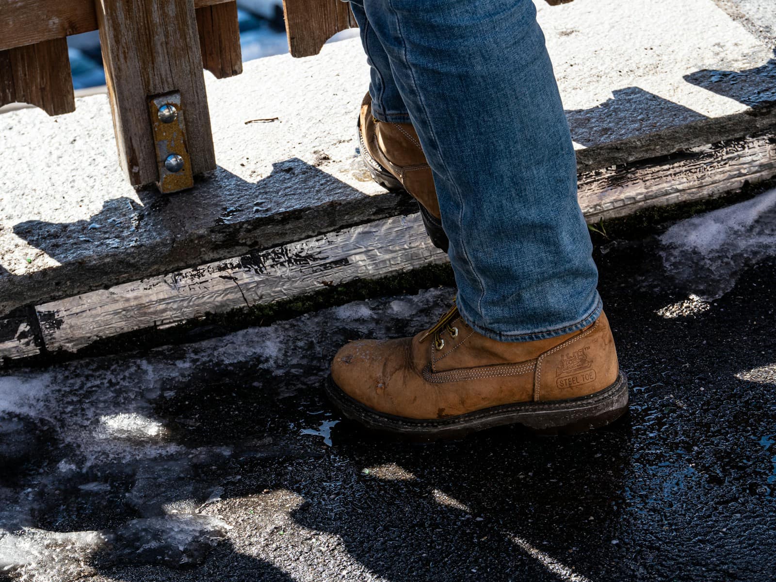 A scuffed leather work boot stepping onto a wet, snow-streaked step, meltwater pooled beside the sole